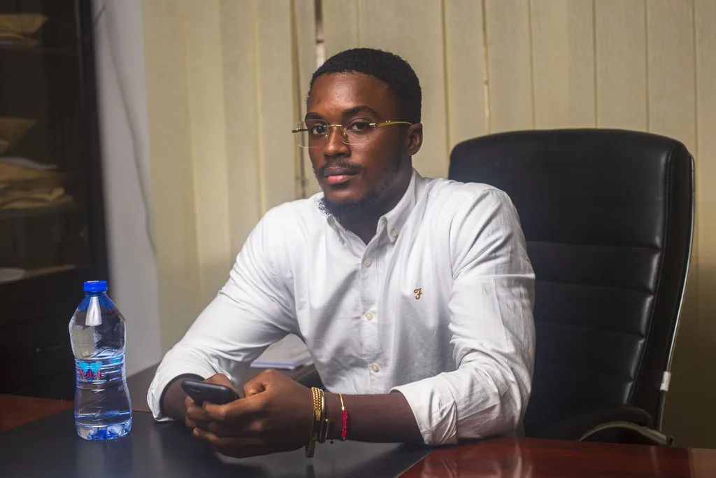 Kwasi Wiafe Kwapong at a desk in a white shirt and rimless glasses, holding a phone and looking into the camera.